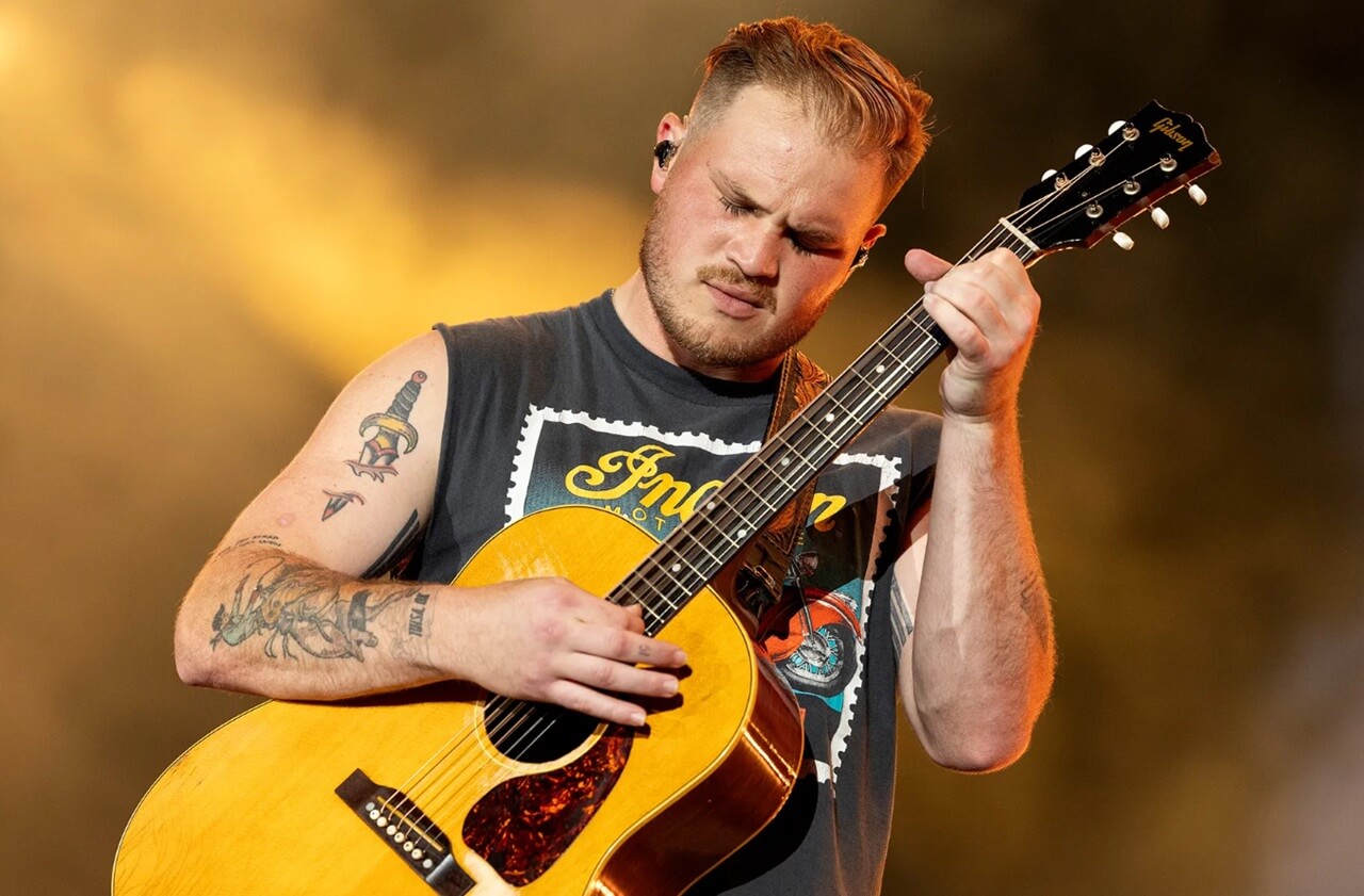 Zach Bryan singing at a concert, holding a guitar, under stage lights.