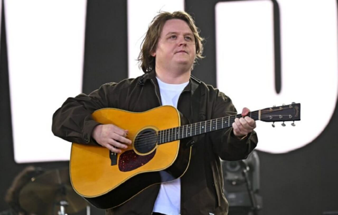 Lewis Capaldi onstage during comeback tour, singing and smiling under bright concert lights.