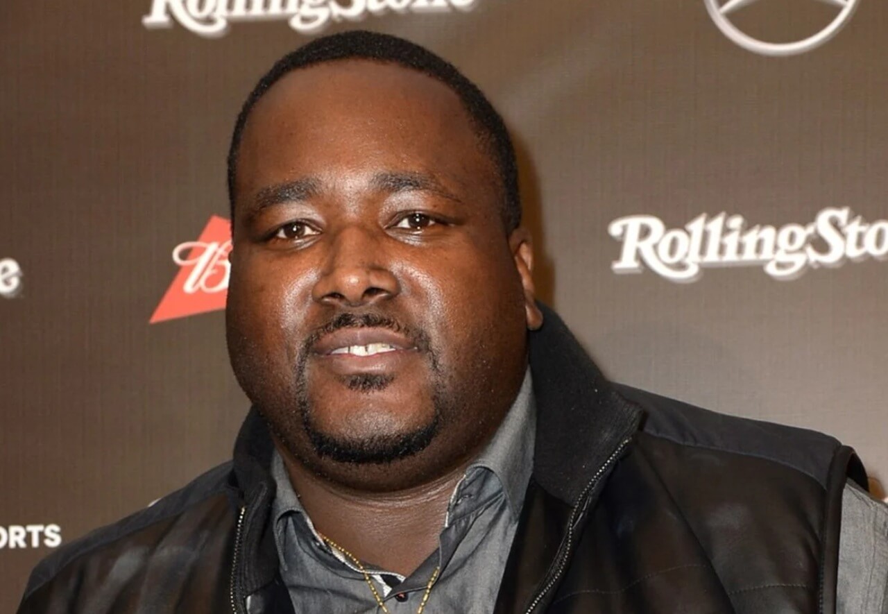 Actor Quinton Aaron attends a red carpet event, smiling in a black suit.