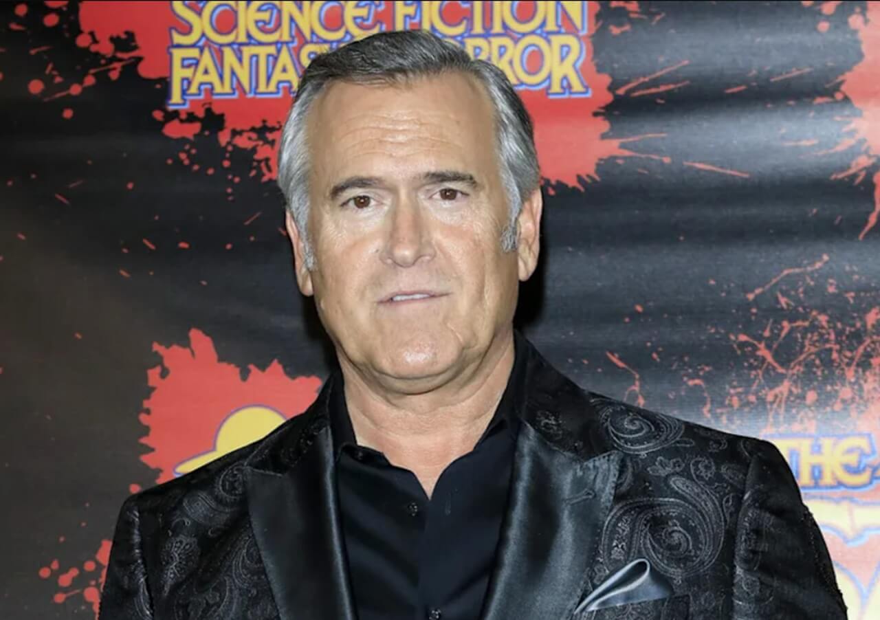 Bruce Campbell at Comic Con, smiling in front of fan art posters.